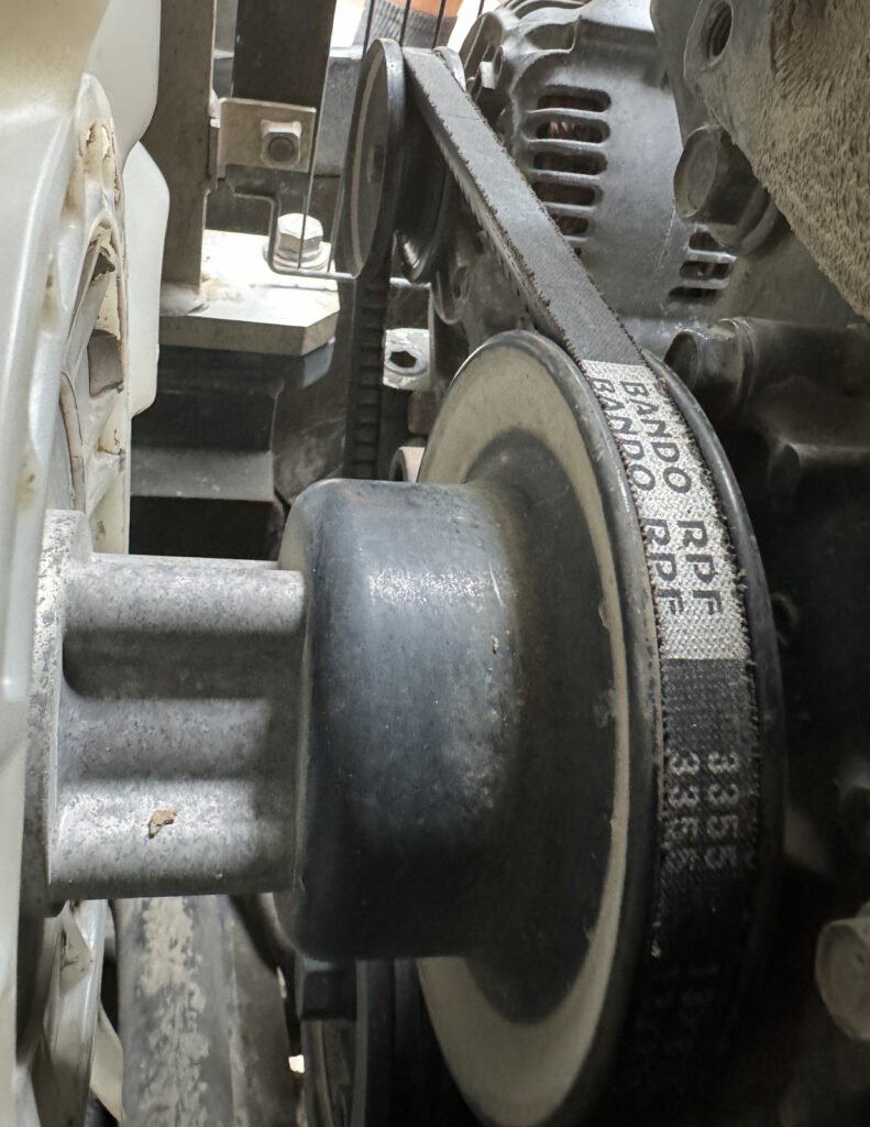 A belt curves around a foreground pulley and runs toward an alternator pulley, with a pale fan blade and metal brackets nearby.