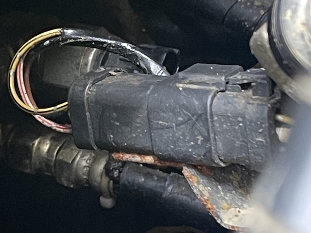 Rounded black connector body beside metalwork, with wires looping from the rear and a locking feature visible near the opposite end.
