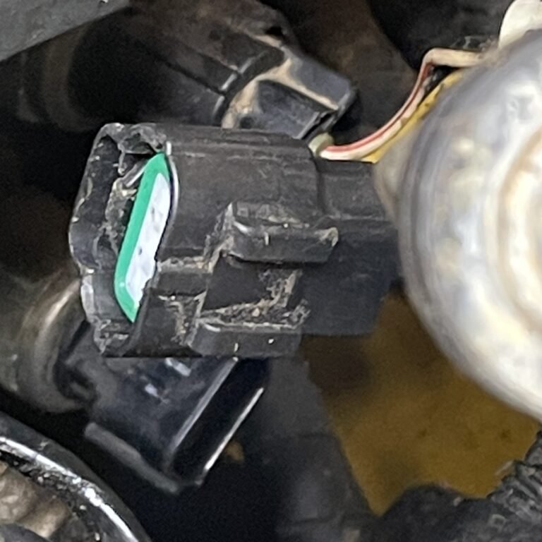 Angled close-up of a black connector with a pale insert, green perimeter, side locking feature and wires visible at the rear.