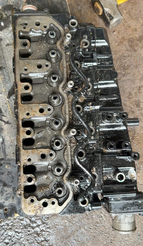 Removed cylinder head with repeated round openings, recessed sections, projecting posts and a narrow dark tube across its open valve area.