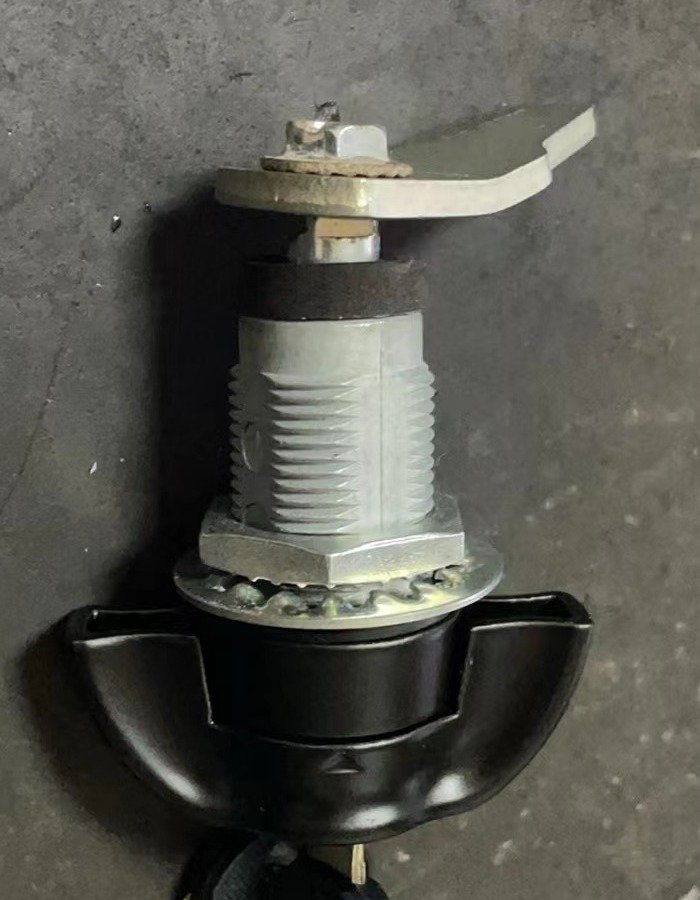 Loose cam lock on a dark surface, showing its black handle, silver threaded body, retaining nut and washer, and offset metal arm.