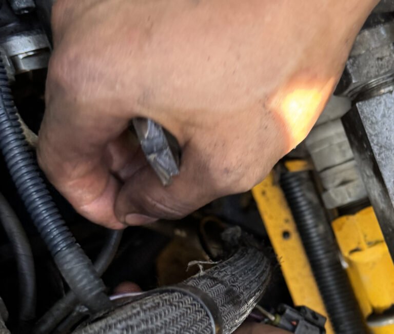 Close photograph of a hand holding a small metal tool beside corrugated sleeving, a fabric-covered line and yellow-painted equipment structure.