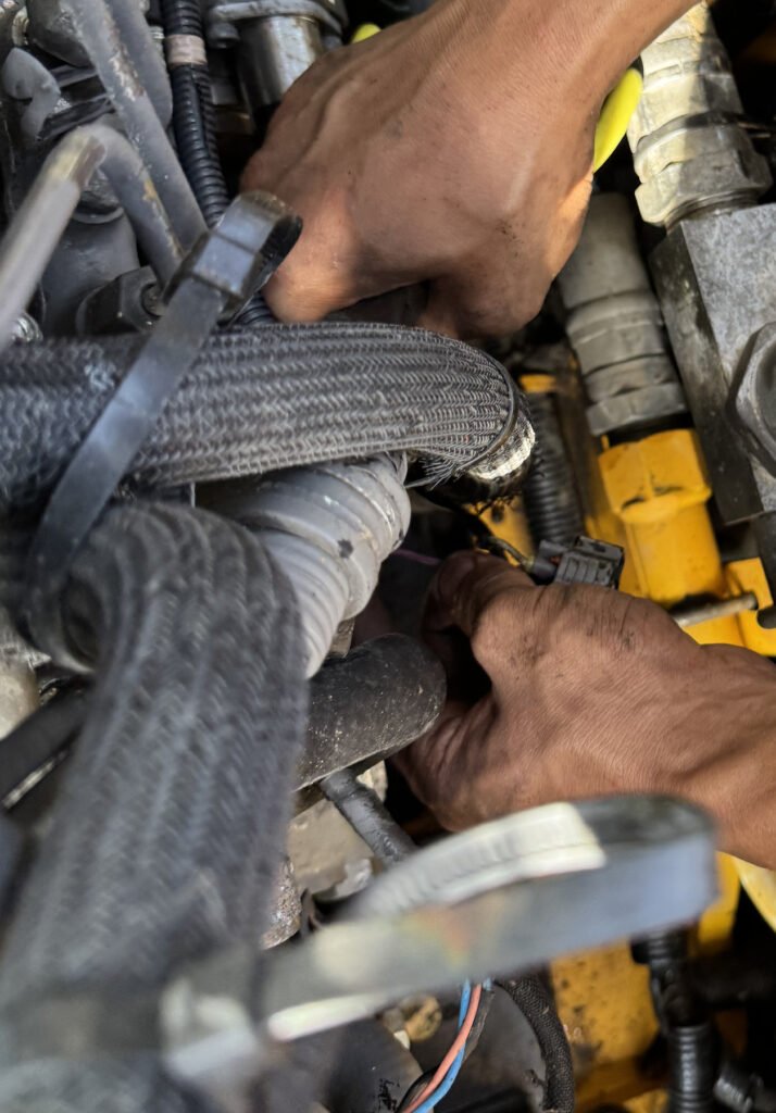 Two hands reaching among overlapping hoses, a small connector and pipework beside yellow-painted equipment structure.