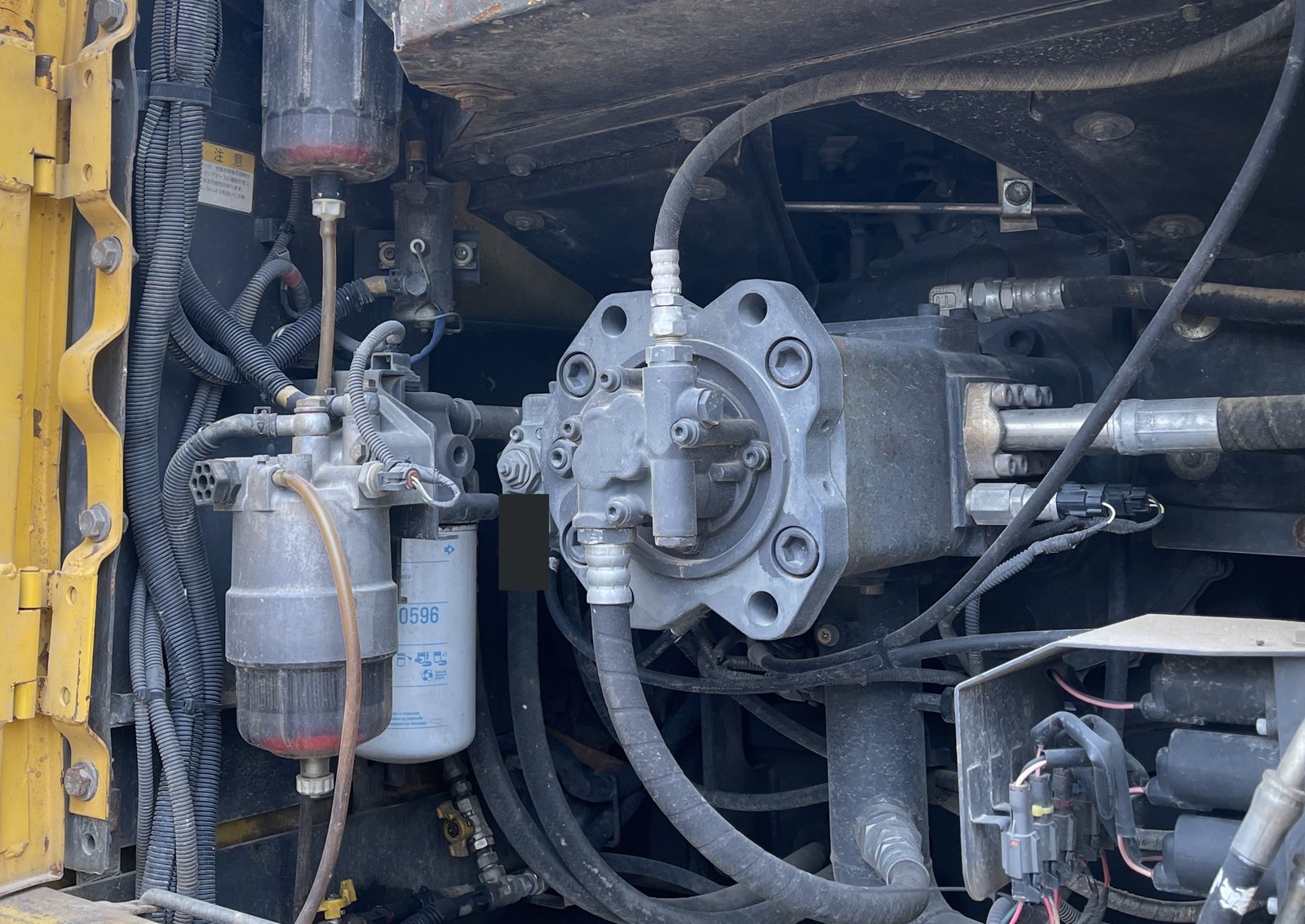 Installed hydraulic-looking assembly with bolted faces, hoses, fittings, filters and wiring nearby.
