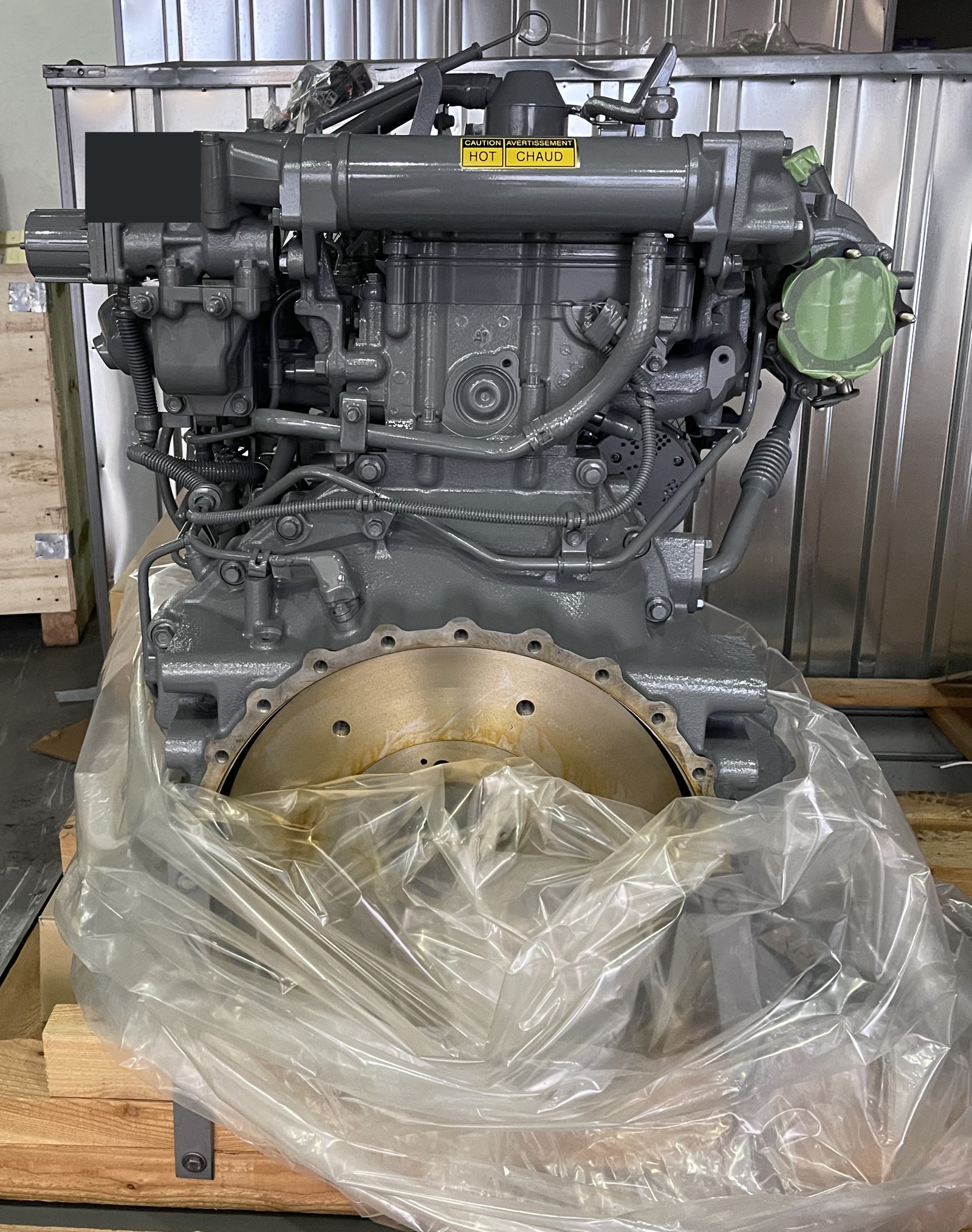 A grey engine assembly on a pallet with protective wrapping, visible accessories and a small upper-left privacy mask.