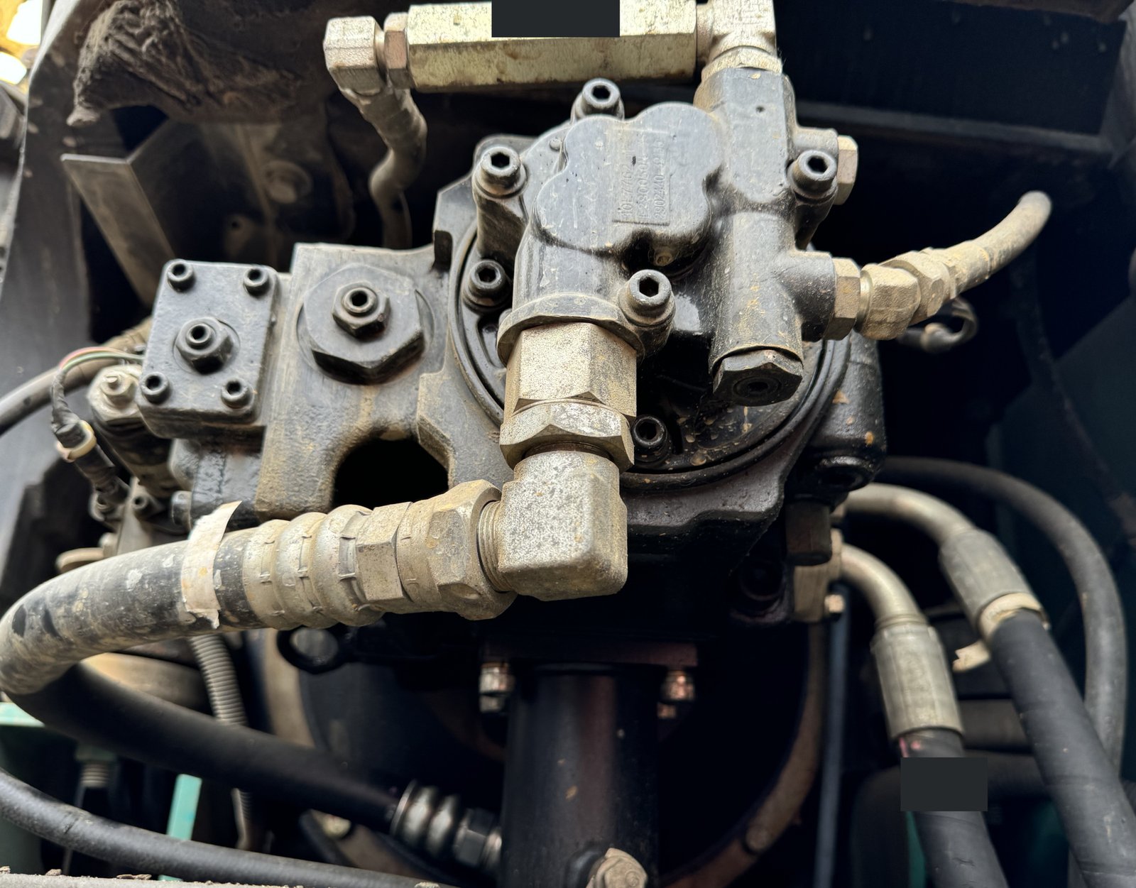 A close installed hydraulic-looking housing with angled fittings, hoses and two small opaque mask areas.