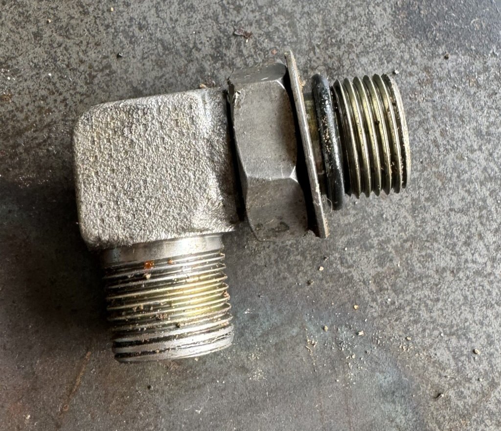 Angled metal fitting with two externally threaded sections, a hexagonal feature and a wider circular feature visible from the side.