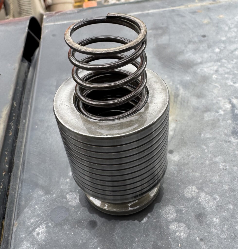 A dark coil spring rises above a circular opening, bright rim and cylindrical metal body with closely spaced outer grooves.
