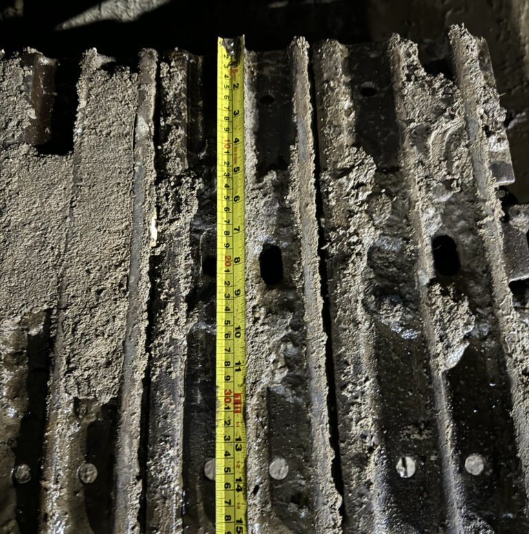 Broad steel track plates with raised ribs, openings, fastener heads and a yellow tape over one plate.